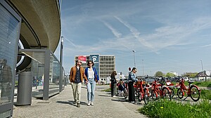 Symbolbild Pärchen läuft auf Weg links Bushaltestelle rechts Fahrradstation und im Hintergrund Parkhaus